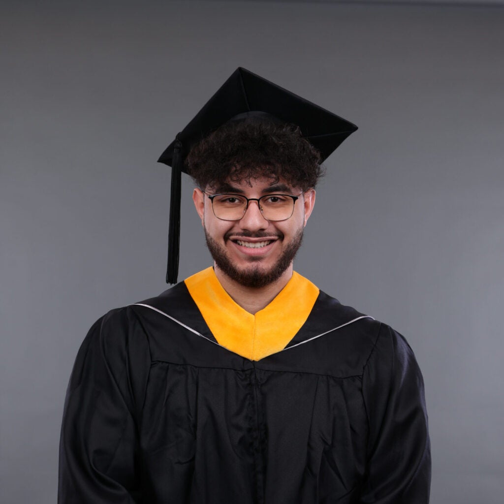 A young man wearing a black graduation gown, cap, and gold stole stands smiling in front of a plain gray background.