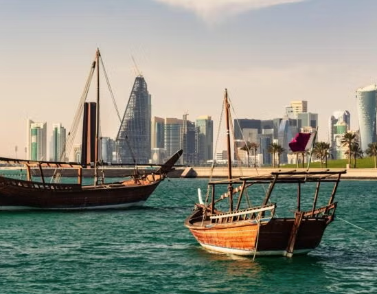 Traditional wooden boats float on turquoise water with a city skyline and modern skyscrapers in the background, under a partly cloudy sky. A maroon and white flag is visible on one of the boats.