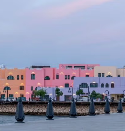 Colorful pastel buildings with arched windows line a waterfront promenade at dusk, with street lamps and a stone railing in the foreground under a soft, clouded sky.