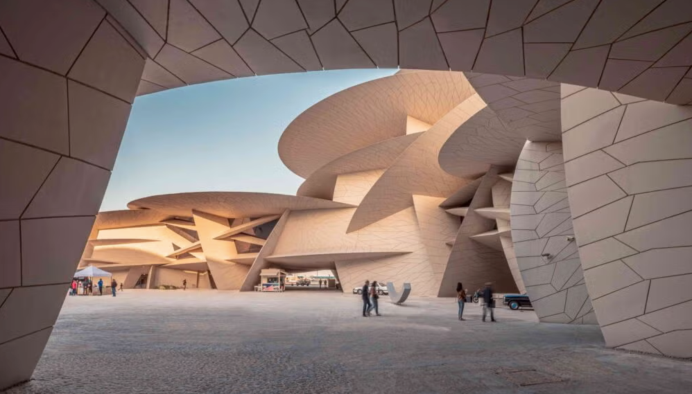 Modern architectural building with interlocking, disc-shaped structures and angular surfaces. Several people walk and stand in the open, sunlit plaza in front of the unique facade.