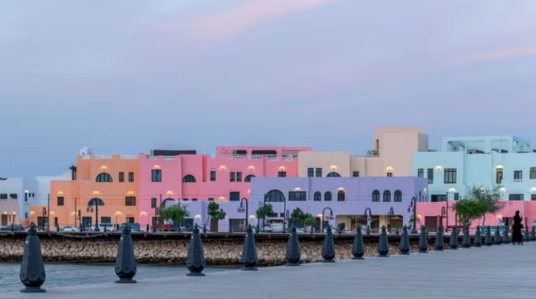A waterfront view of pastel-colored buildings in shades of pink, purple, blue, and beige, with a row of black bollards along a promenade and a calm sky at dusk.
