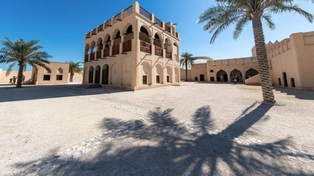 A traditional beige fort with arched windows and palm trees casting shadows stands under a clear blue sky in a sandy courtyard.