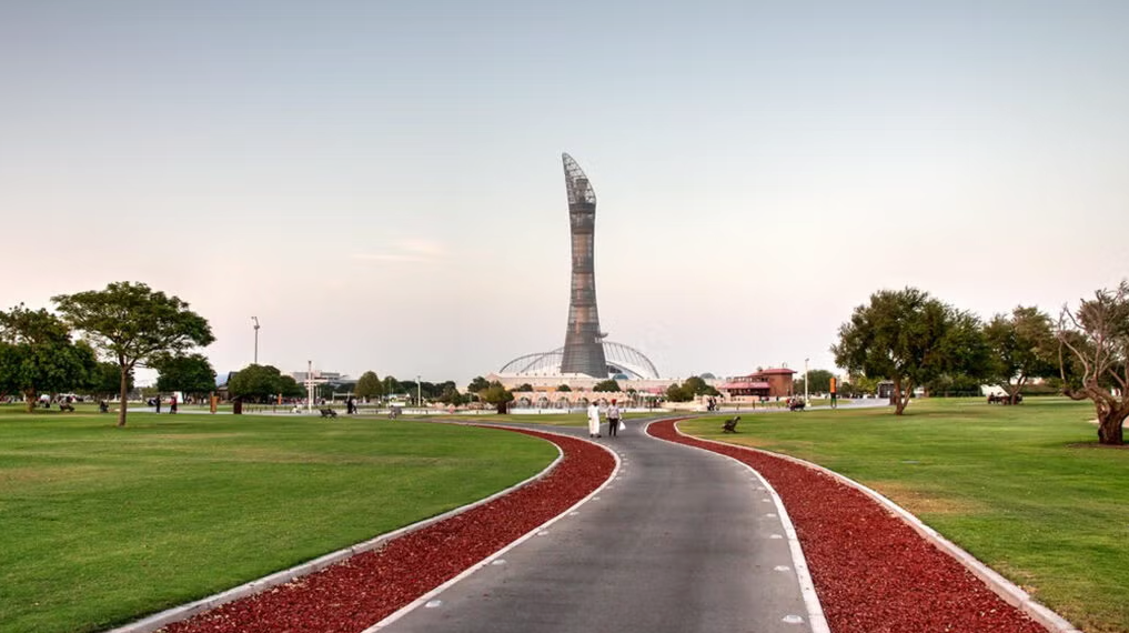 A paved walkway with red trim curves through a green park toward a tall, uniquely shaped tower under a clear sky; people are walking along the path, and trees are scattered on the grass.