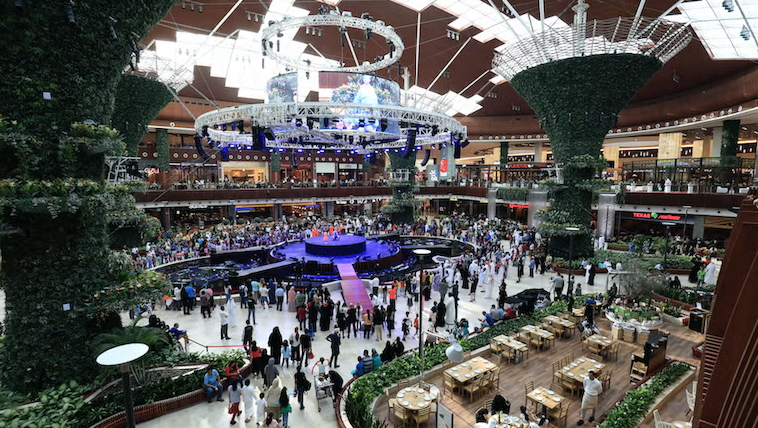 A large indoor mall atrium with greenery, a central stage, and crowds of people gathered around. There are hanging lights, surrounding shops, and dining areas filled with tables and chairs.
