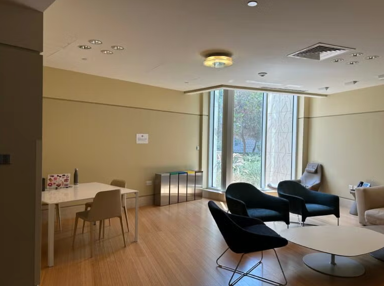 A modern lounge area with light wood floors, several black chairs around a round white table, a window with natural light, and a small table with beige chairs on the left side.