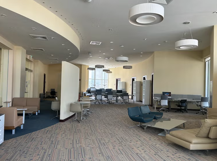 A modern, spacious office lounge with various seating areas, including chairs, sofas, and desks with computers. Large windows let in natural light, and circular ceiling lights hang above. The decor features neutral colors.