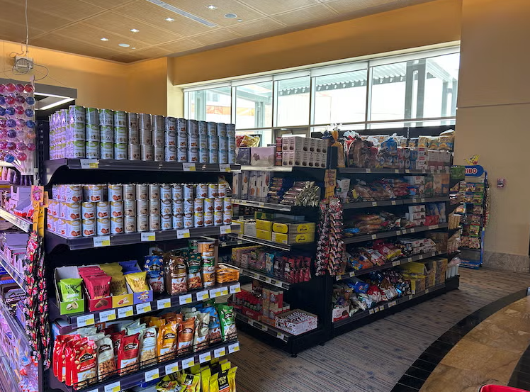 Shelves stocked with canned goods, snacks, and packaged foods inside a brightly lit convenience store. Sunlight streams through large windows, illuminating the aisles and displays.