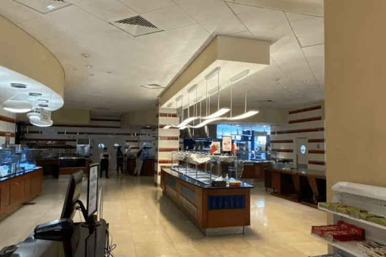 A spacious, modern cafeteria with buffet stations, bright overhead lighting, and a few people serving food in the background. The floor is tiled and the walls have horizontal striped accents.