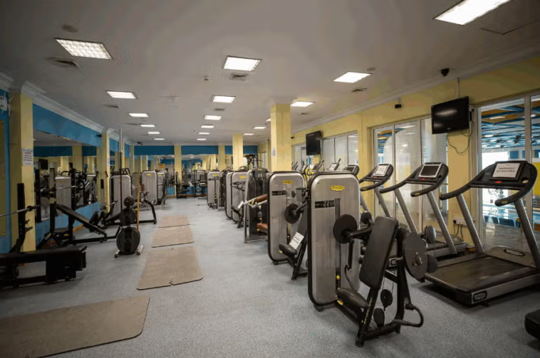 A spacious gym with various fitness machines, including treadmills, weight benches, and strength-training equipment. Large mirrors line the walls, and bright overhead lights illuminate the area.