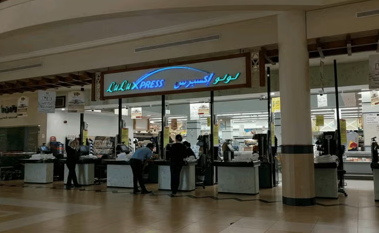 A supermarket checkout area with several counters, some staffed by employees and some customers waiting. Above is a lit sign reading Lulu Xpress in English and Arabic. The interior is well-lit, while the mall outside is dimmer.