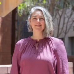 A person with shoulder-length gray hair stands outdoors, wearing a long-sleeved, mauve blouse. Trees and buildings are visible in the background.