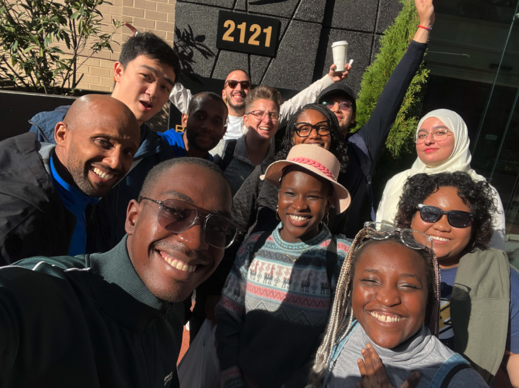 A diverse group of people are smiling and posing for a selfie outside, in bright sunlight, in front of a building with the number 2121 displayed above the entrance.