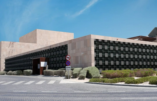 A modern, angular building with beige stone walls and a grid-like black facade, set against a blue sky. There are trimmed bushes, road signs, and a crosswalk in the foreground.