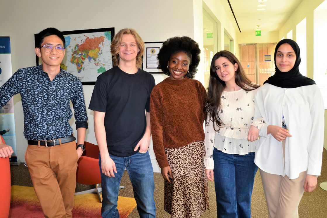 Five young adults stand together indoors, smiling at the camera. They are casually dressed and represent diverse backgrounds. Behind them are maps on the wall and a hallway with natural light.