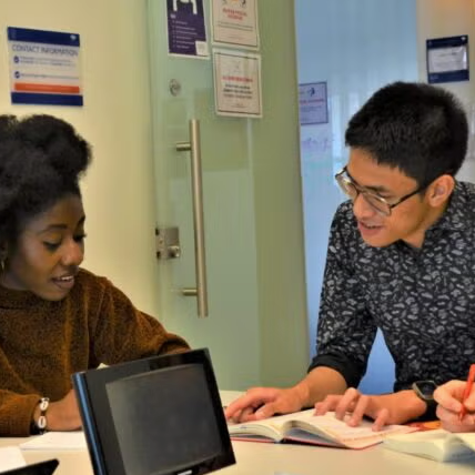 Three students sit at a table in a bright room, collaborating over open books and notebooks. One student points to a page, while the others listen and take notes. Signs and notices are visible on the door behind them.