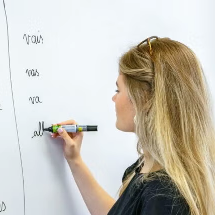 A woman writes French verbs on a whiteboard, listing subject pronouns and forms of aller (to go). She holds a marker and faces the board, with her long blonde hair down and sunglasses on her head.