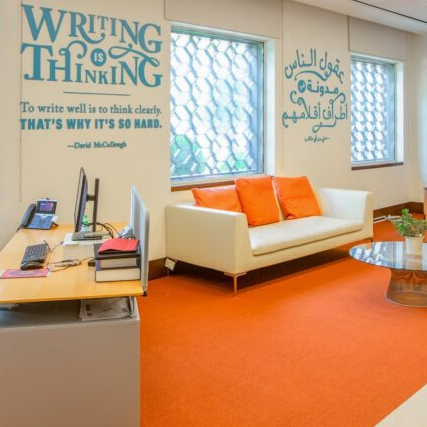 A bright office with orange carpet, a white sofa with orange pillows, a glass coffee table, desks with computers, bookshelves, and inspirational quotes on the walls in English and Arabic. Large windows let in natural light.