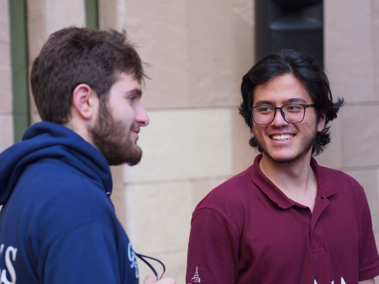 Two young men stand outside, talking and smiling at each other. One wears a blue hoodie and has a beard, while the other wears glasses and a maroon polo shirt. Cream and beige tiles make up the background wall.