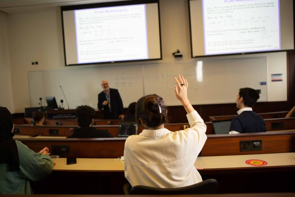 A student raises their hand to ask a question in a university classroom while a professor stands at the front near a presentation screen. Other students are seated, listening and taking notes.
