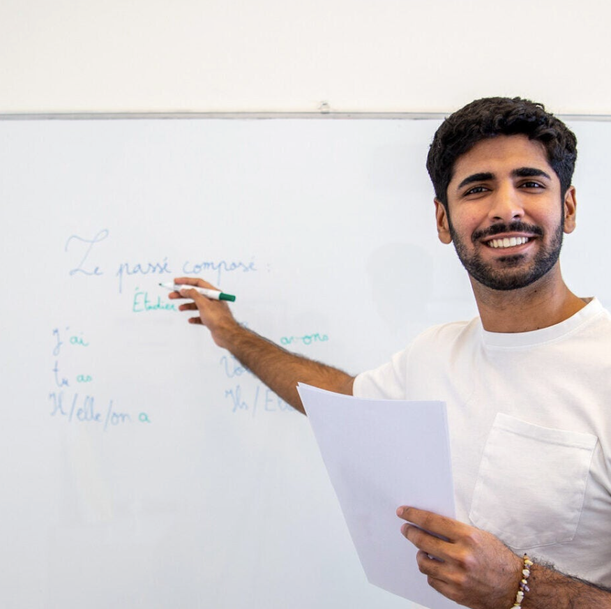 A man holding papers smiles while pointing at a whiteboard with French writing on it, suggesting he is teaching or giving a presentation.