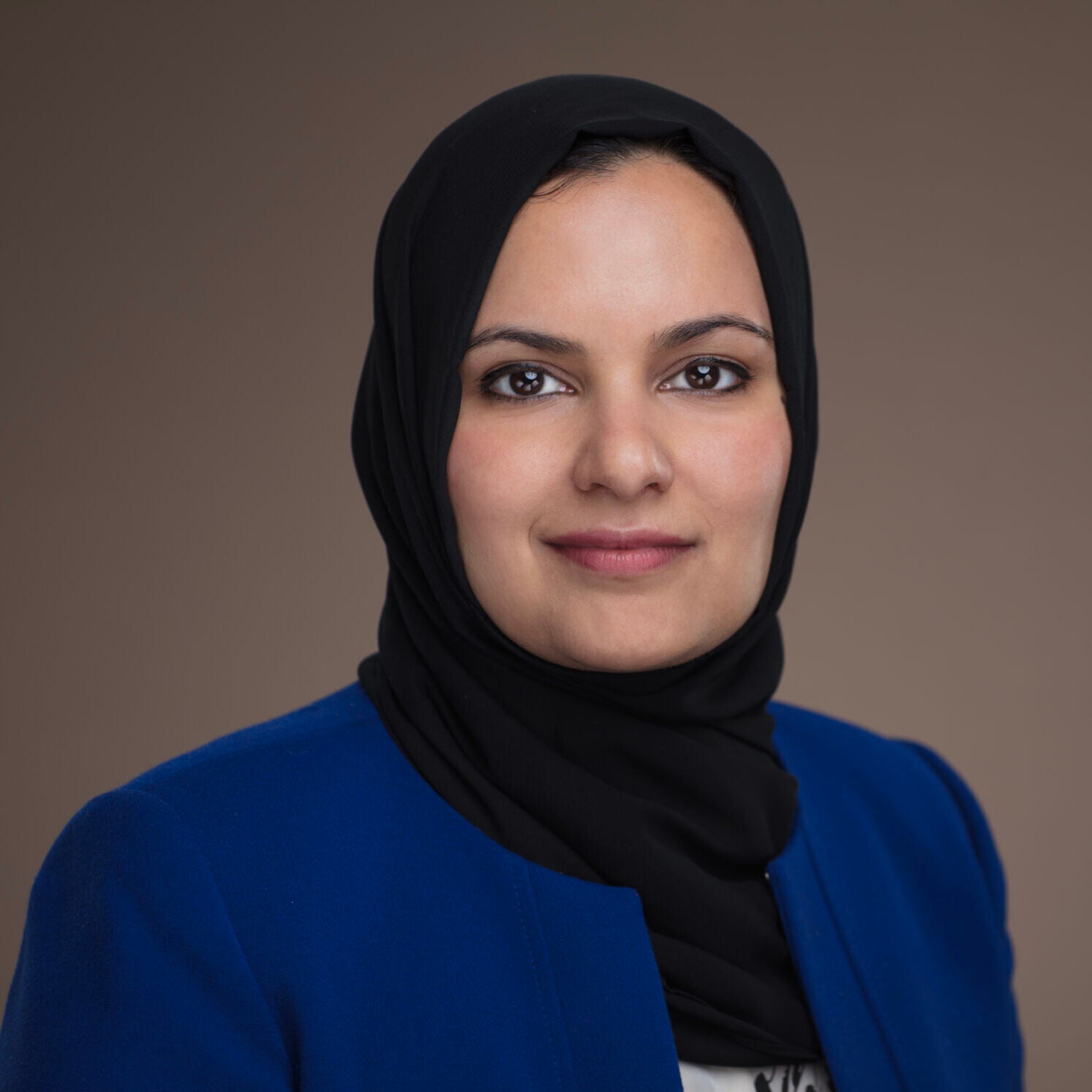 A woman wearing a black hijab and a blue blazer faces the camera and smiles slightly, posing against a plain brown background.