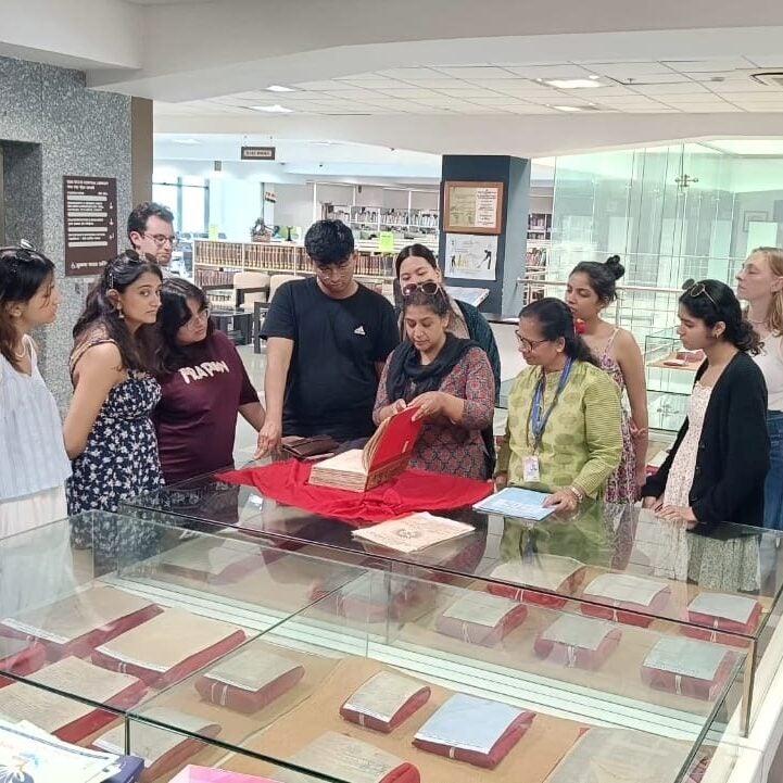 A group of people stand around a glass display case in a library or museum, listening to a woman who is presenting an open book placed on a red cloth, with several other books visible in the case.