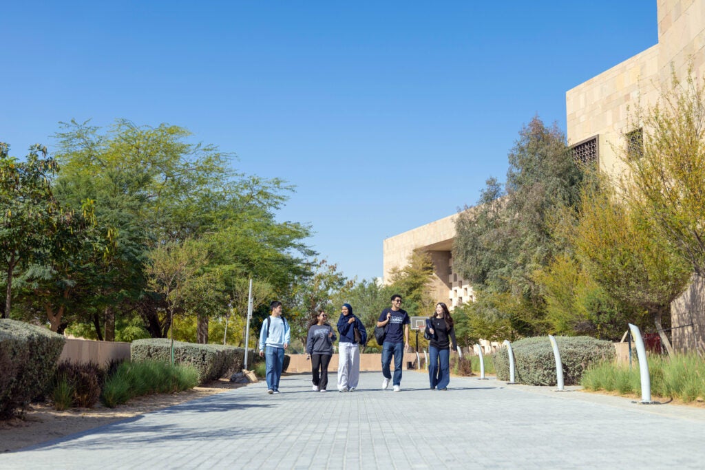 Five students walk together on a paved pathway lined with greenery and outside the modern tan GU-Q building under a clear blue sky, appearing relaxed and engaged with each other.
