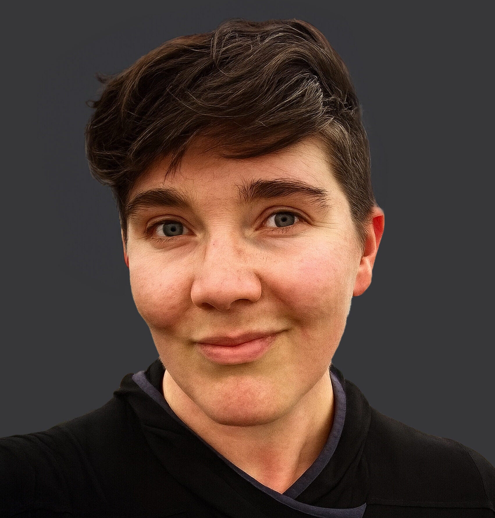 A person with short brown hair and blue eyes, wearing a black top, smiles slightly at the camera against a dark, plain background.
