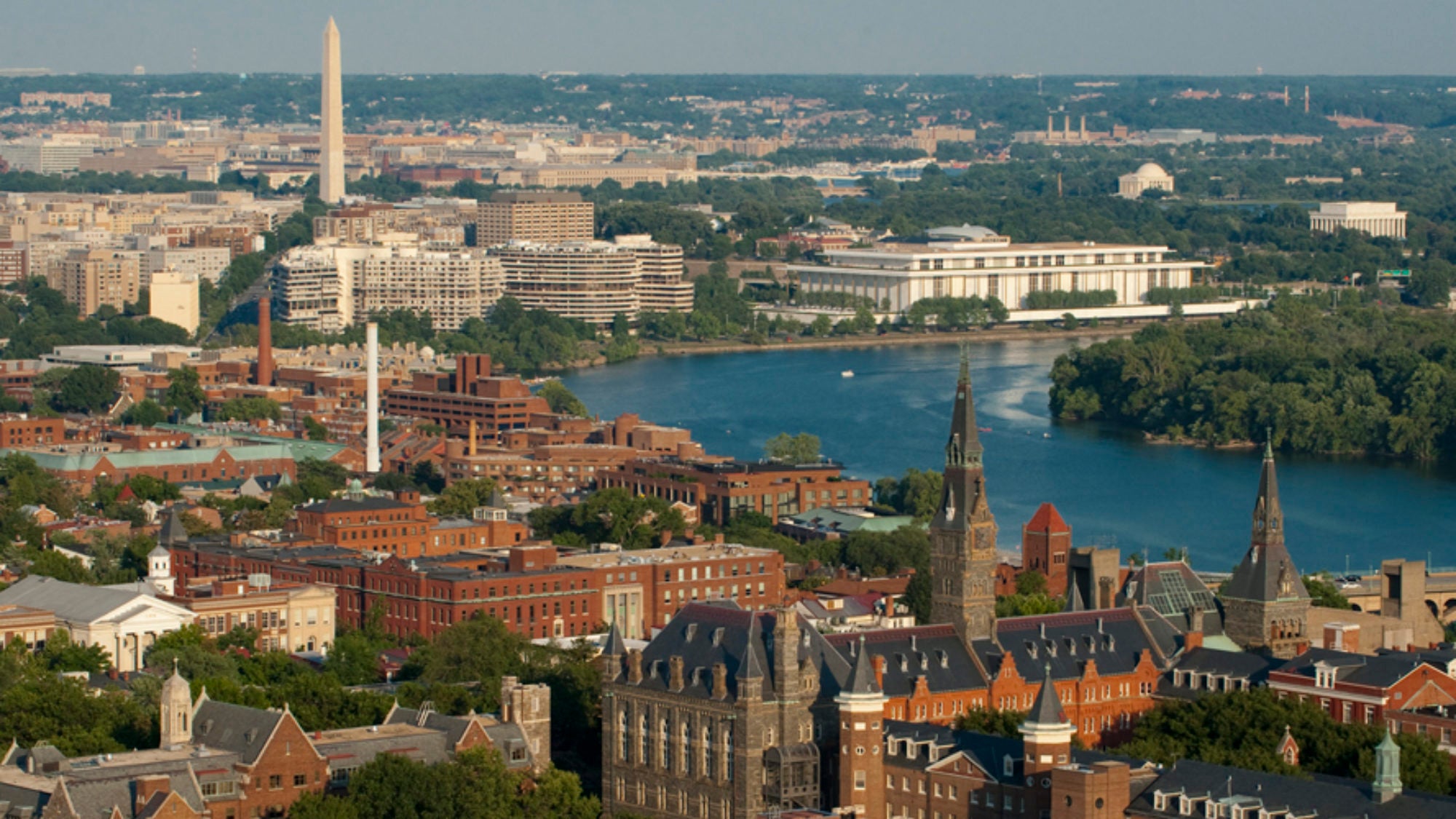 Aerial view of Georgetown University’s campus in Washington, D.C., with historic buildings in the foreground, the Potomac River, and city landmarks including the Washington Monument and the Kennedy Center in the background.
