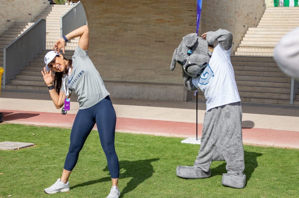 A woman in exercise clothes stands on a field stretching. Georgetown's mascot, Jack the bulldog, is standing next to her stretching in the same way.