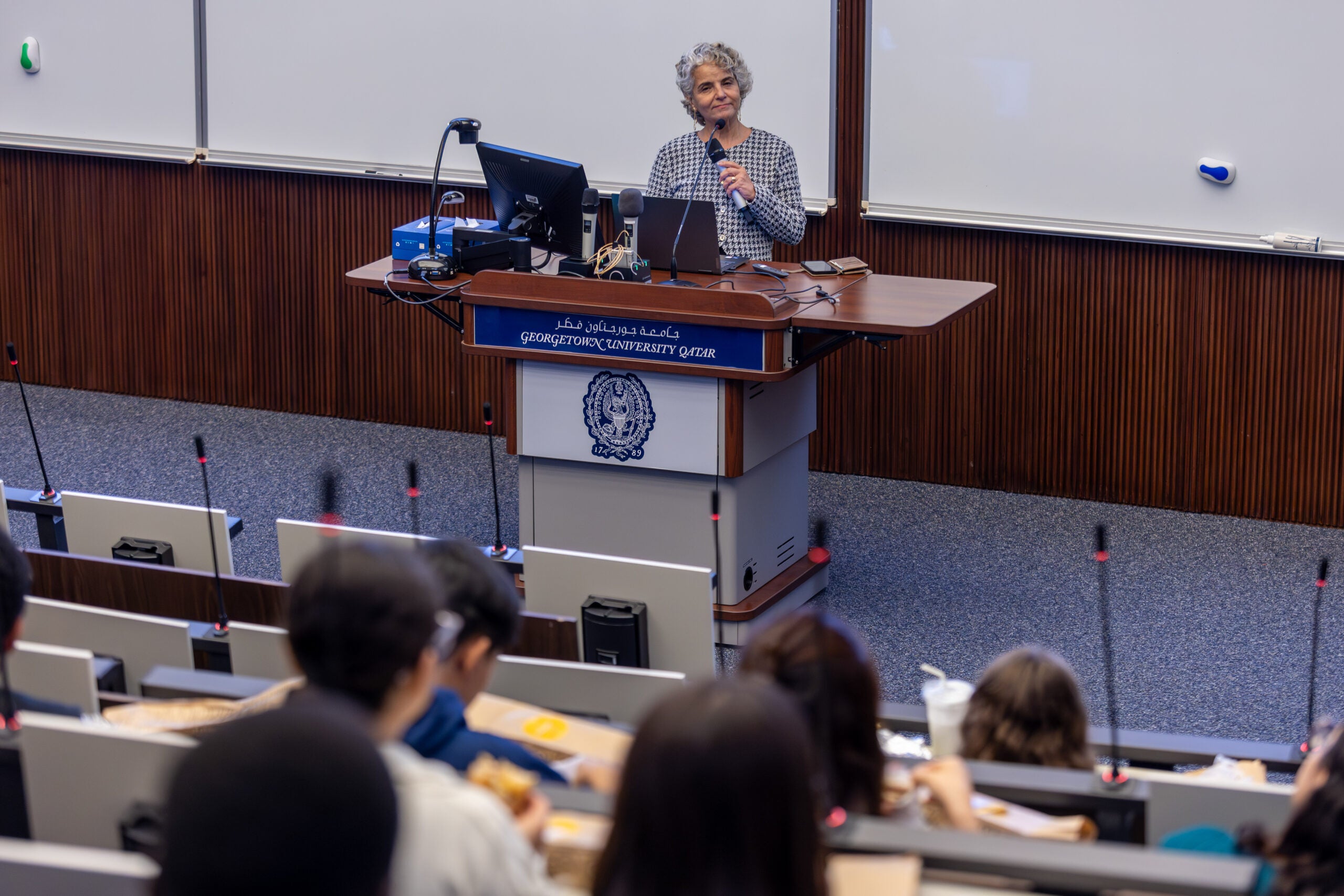 A woman stands behind a podium in an auditorium speaking to a group of assembled students.