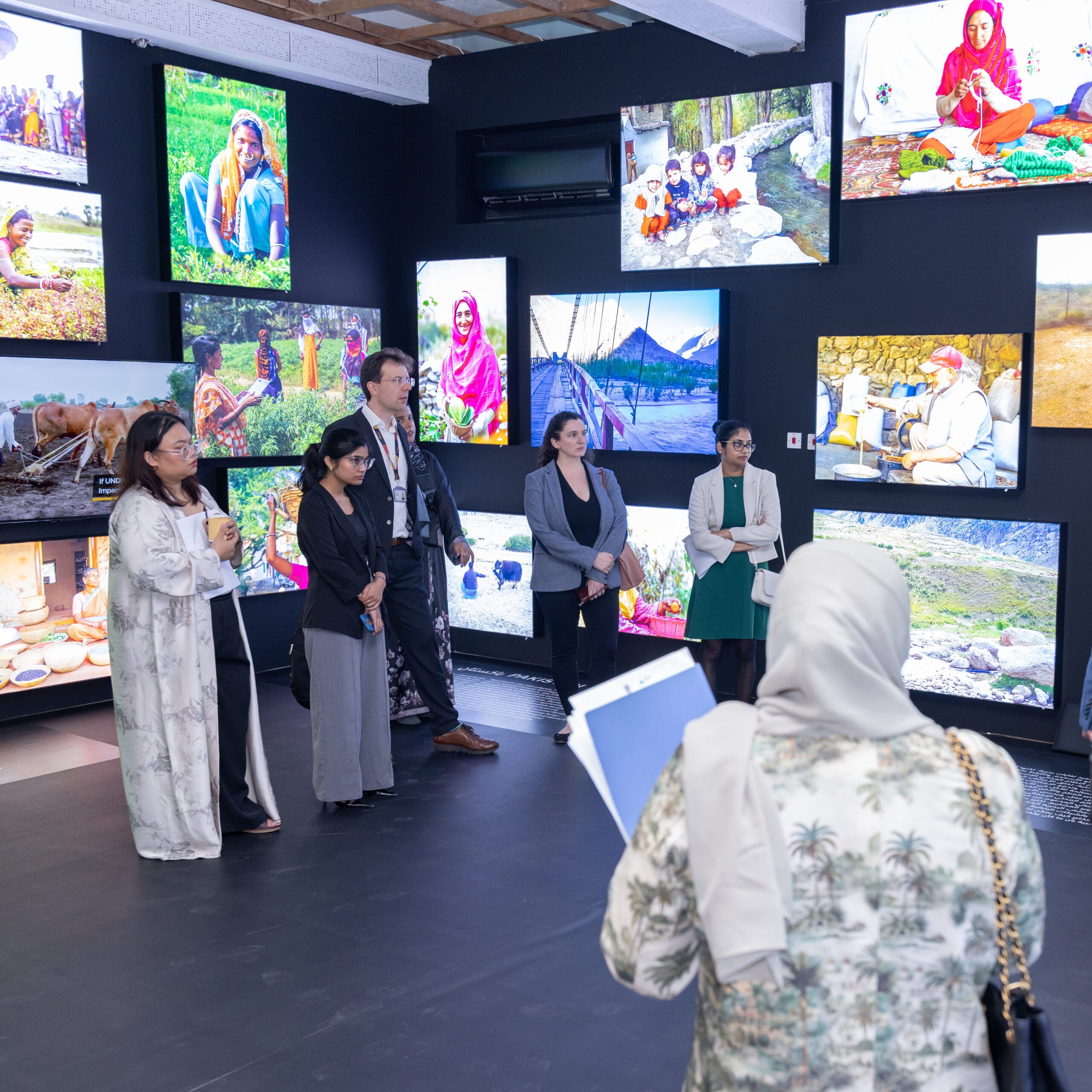 A group of people in formal attire stands in a gallery room, listening attentively, surrounded by vibrant photographs of diverse people and landscapes displayed on the walls.