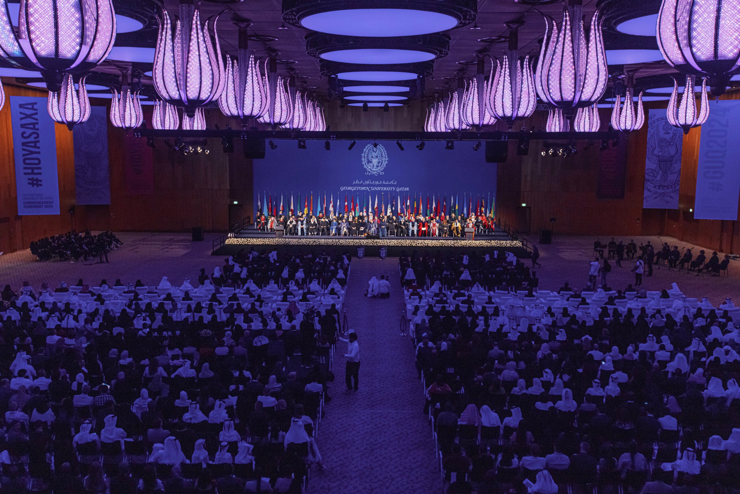 A large conference hall filled with seated attendees faces a stage with panelists and many international flags; purple lighting creates a formal atmosphere.