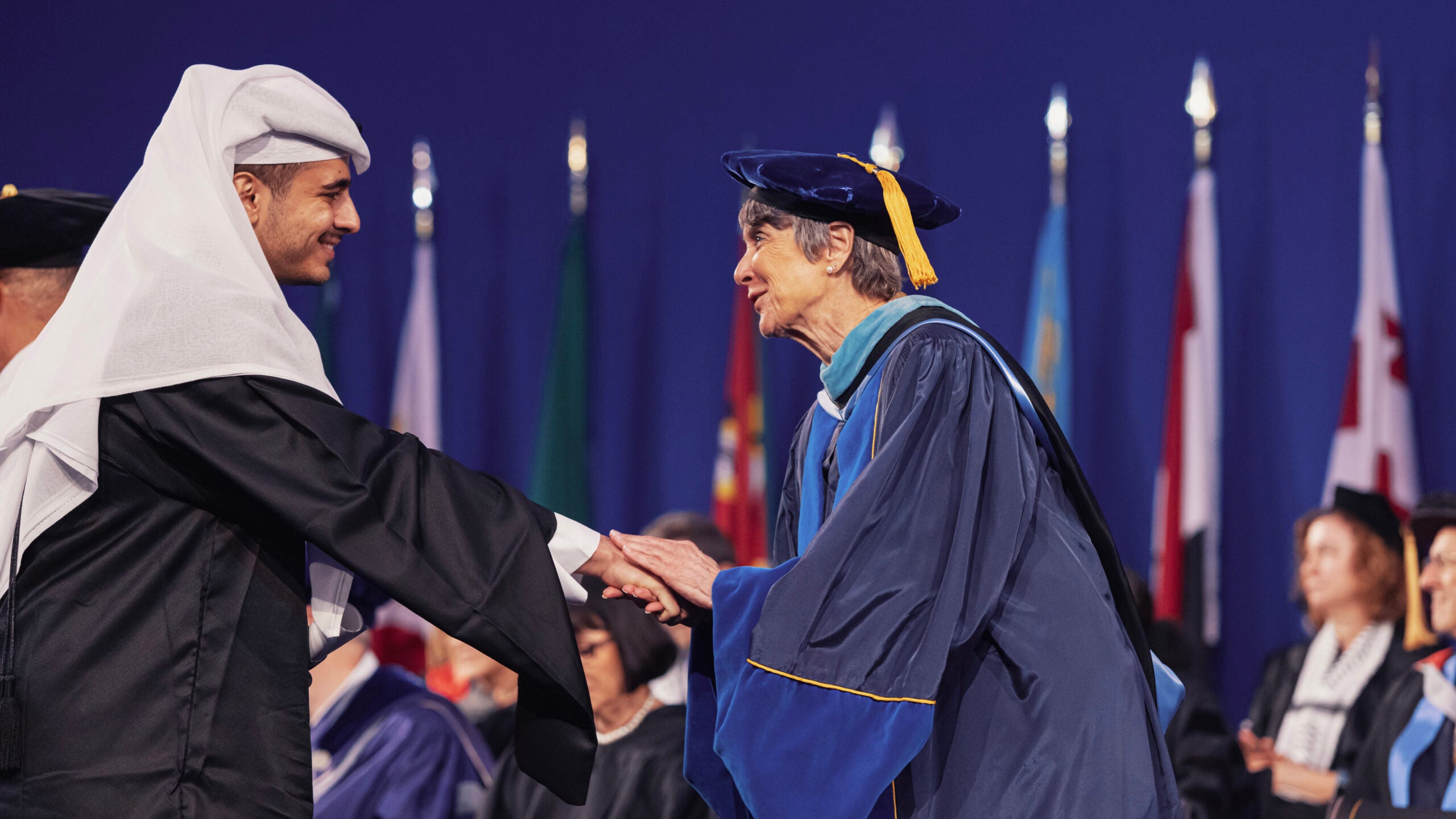 A graduate from the master's program in traditional attire shakes hands with a faculty member in academic regalia on stage at a graduation ceremony, with international flags in the background.