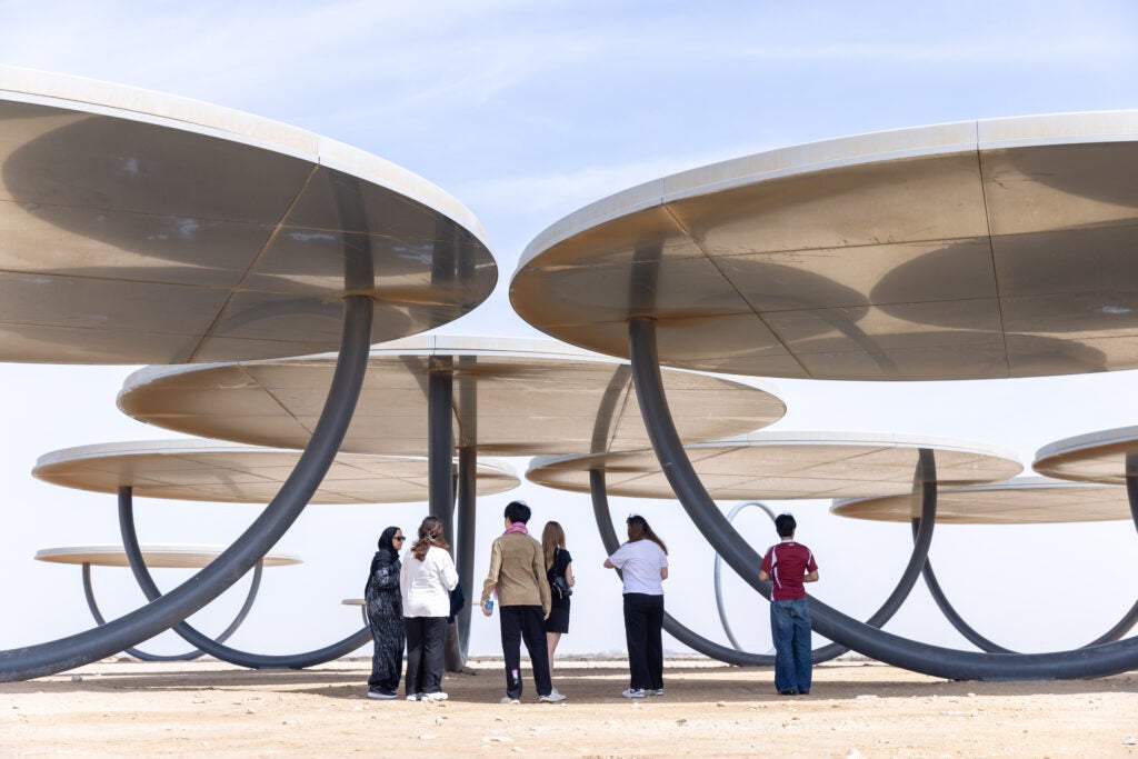 Six people stand under large, futuristic, disc-shaped metal structures that provide shade on a sandy ground under a partly cloudy sky.
