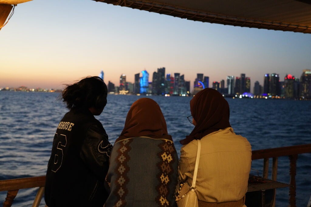 Three people sit on a boat at sunset, looking toward a city skyline across the water, with illuminated buildings in the distance.