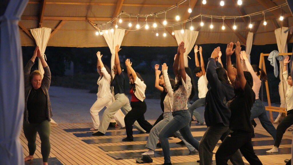 A group of students do yoga poses under a canopy with warm, yellow lights strung above them.