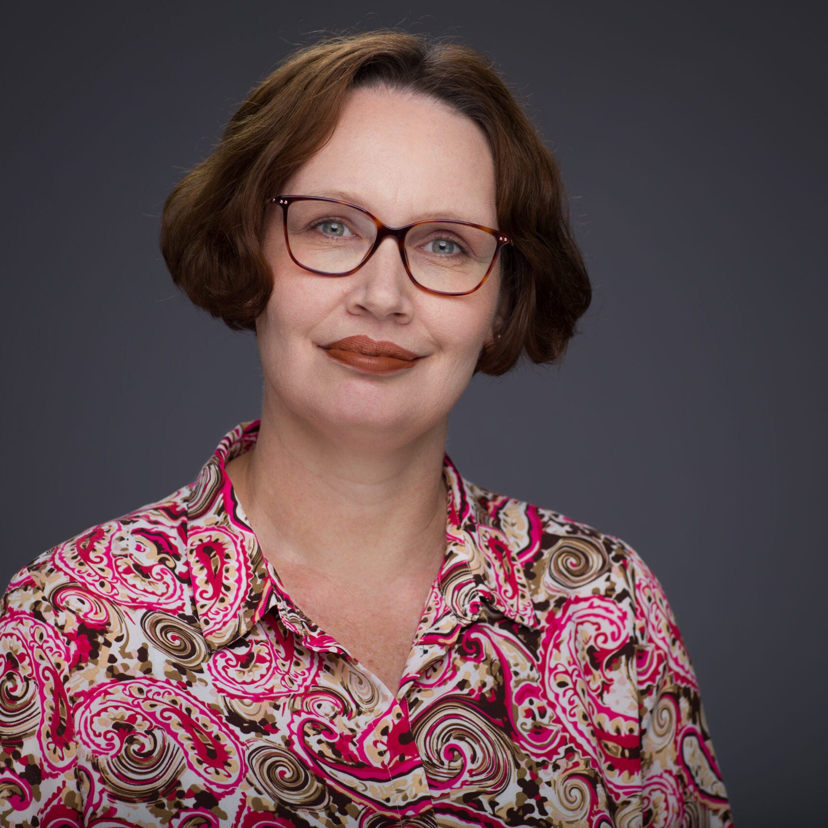 A woman with short brown hair, wearing glasses and a pink paisley-patterned blouse, smiles softly against a plain dark gray background.