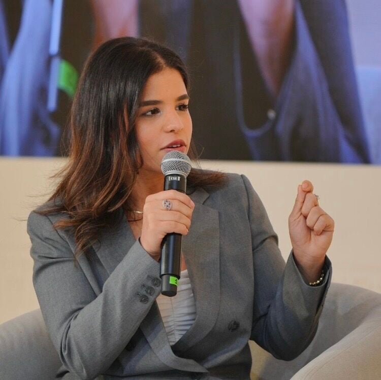 A woman in a gray suit sits in a chair, holding a microphone and speaking, gesturing with her hand. She appears engaged in conversation, with a blurred background behind her.