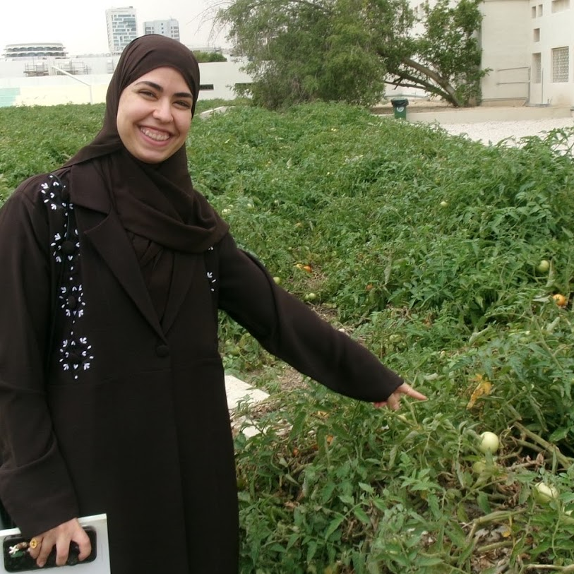 A smiling woman wearing a brown hijab and long coat stands next to a lush green garden bed, pointing at growing tomato plants. Urban buildings are visible in the background.