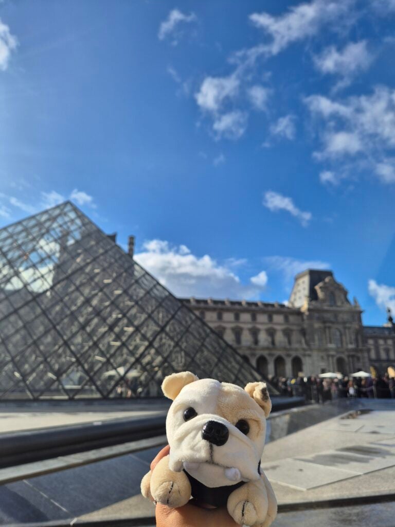 A hand holds a plush bulldog toy in front of the Louvre Pyramid in Paris, with blue sky and scattered clouds in the background.