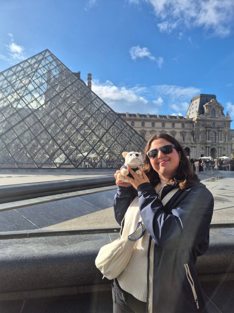 A woman wearing sunglasses and a navy jacket smiles while holding a stuffed animal in front of the Louvre Pyramid in Paris on a sunny day.