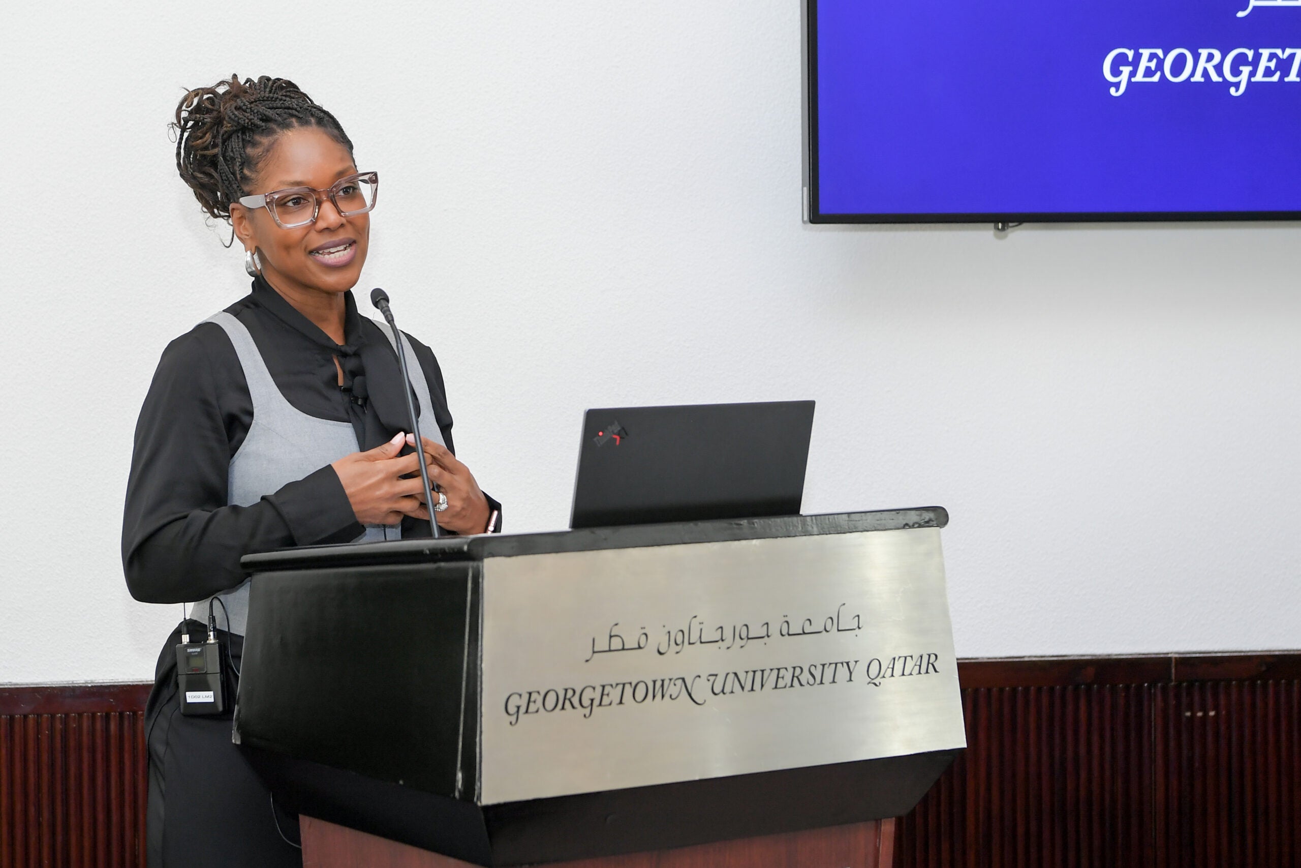 A woman stands at a podium labeled Georgetown University Qatar, speaking with a microphone and gesturing with one hand, while a screen with the university logo is visible in the background.