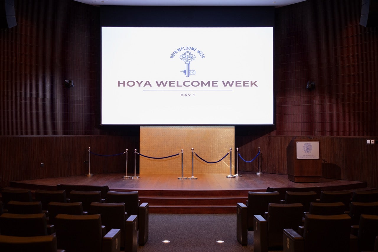An empty auditorium with rows of chairs faces a stage and a large screen displaying HOYA WELCOME WEEK DAY 1. A podium stands to the right, and a blue rope barrier is in front of the stage.