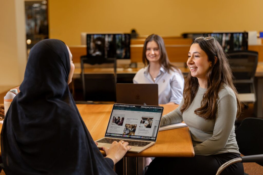 Three women sit at a table in a library or office, talking and working on laptops. One wears a hijab, another wears a grey top and smiles, and the third sits in the background with a laptop.
