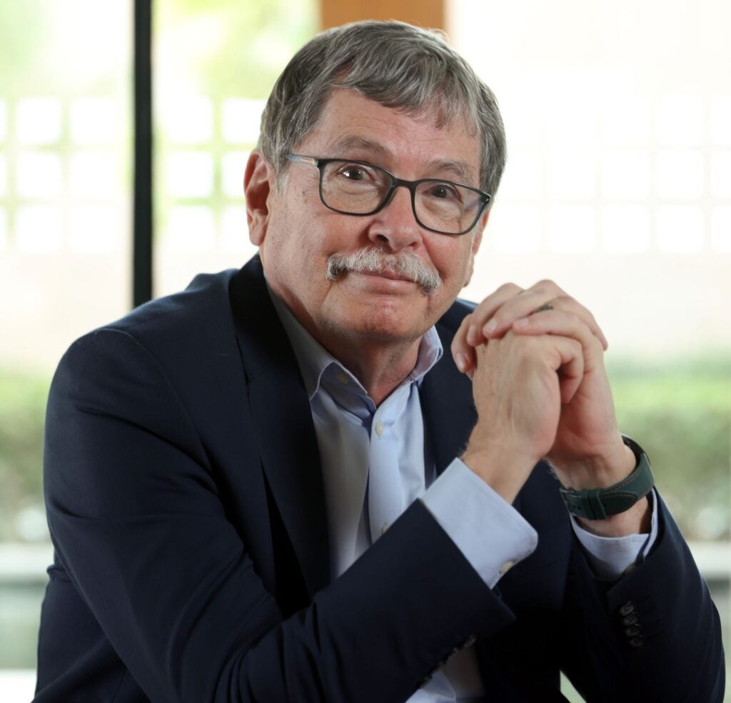 An older man with gray hair, glasses, and a mustache, wearing a dark blazer and light blue shirt, sits indoors with hands clasped and a window with blurred greenery in the background.