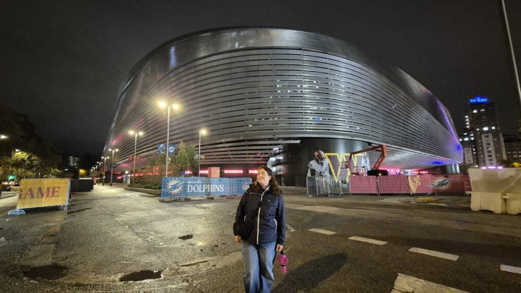 A person stands on a wet street at night in front of a large, modern, curved building with lights. Construction barriers and equipment are visible around the area.