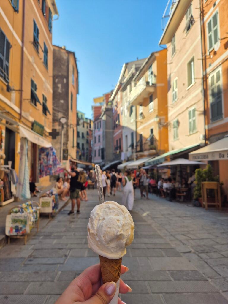 A hand holds an ice cream cone in a lively street lined with colorful buildings, shops, and outdoor cafes under a clear blue sky. People are walking and sitting at tables in the background.