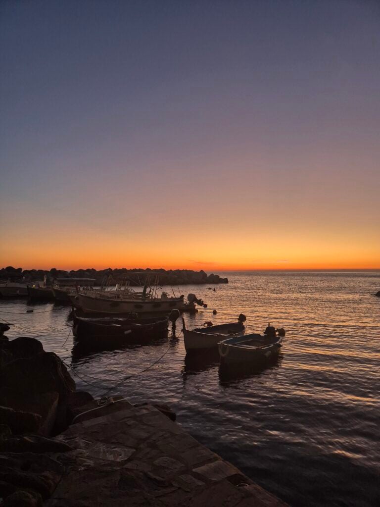 Small boats float on calm water at sunset, with an orange and purple sky, silhouetted boats, and a rocky shoreline in the foreground.
