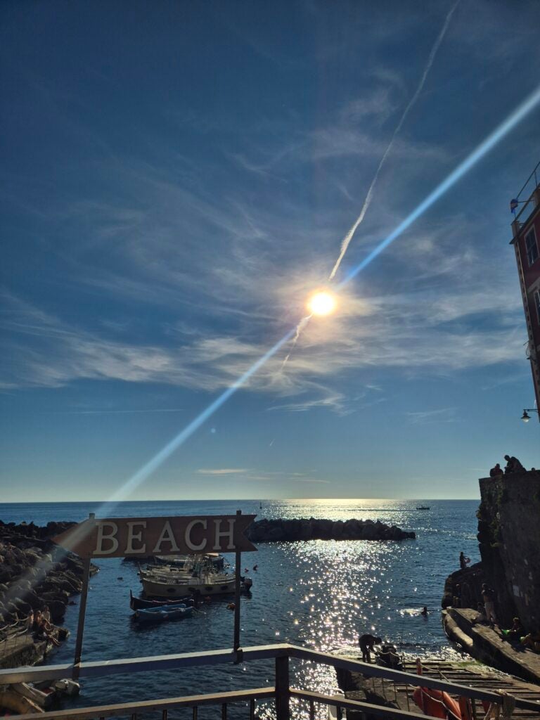 Sun shines over a coastal scene with boats on the water, a BEACH sign in the foreground, and silhouettes of people near the shore. Blue sky with light clouds and a building at the right edge.